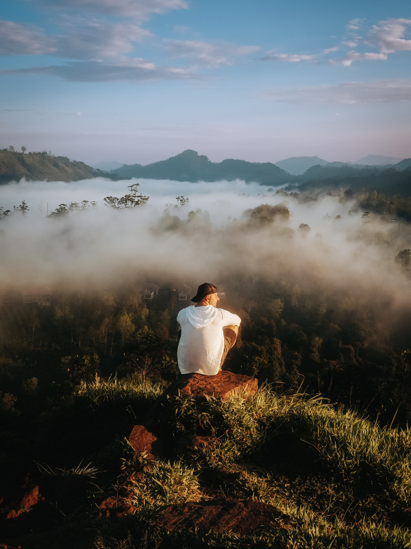 Widok z Little Adam’s Peak na Sri Lance o wschodzie słońca, turysta siedzący na skale nad morzem mgieł i zielonymi wzgórzami