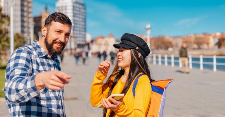 dwóch ludzi na świeżym powietrzu, w słoneczny dzień, na tle miejskiego krajobrazu. Po lewej stronie znajduje się mężczyzna z krótką brodą i w kraciastej koszuli, który pokazuje coś palcem w oddali. Po prawej stronie jest uśmiechnięta kobieta w czarnym kaszkiecie i żółtej bluzie, trzymająca telefon komórkowy. W tle widać ludzi spacerujących po deptaku oraz budynki miasta. [s] shutterstock