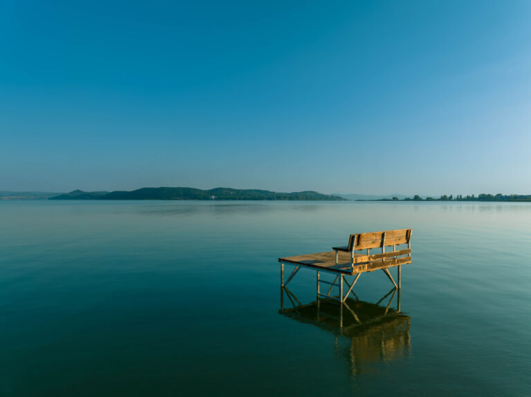 Drewniana ławka na niewielkim molo znajdującym się na spokojnej tafli jeziora Balaton, z widokiem na Tihany półwysep w tle. Błękitne niebo i spokojna woda tworzą spokojny, relaksujący krajobraz.