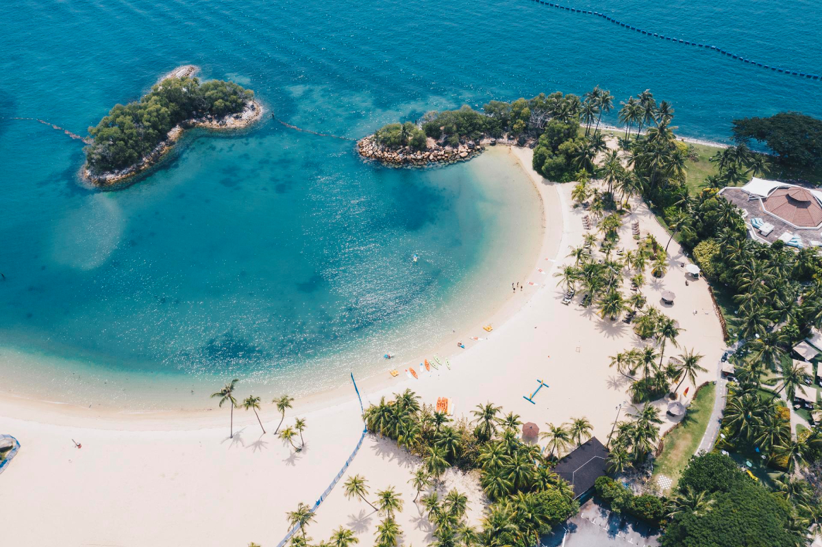 Sinagpur. Sentosa Island. Widok z lotu ptaka przedstawia plażę z białym piaskiem i turkusową wodą. Wzdłuż linii brzegowej rosną palmy. Na wodzie znajdują się dwie małe wyspy porośnięte zieloną roślinnością. Po prawej stronie zdjęcia widoczny jest budynek z brązowym dachem.