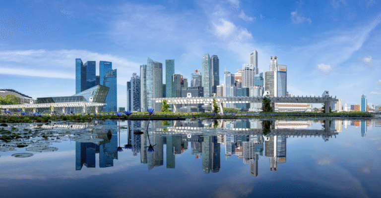 Panoramiczny widok miasta Singapur odbija się w spokojnej tafli wody, a nad nim rozciąga się czyste, błękitne niebo. Horyzont tworzą wieżowce o różnej wysokości i konstrukcji. Na pierwszym planie tafla wody odbija budynki i niebo. Po lewej stronie widoczna jest nowoczesna konstrukcja architektoniczna z szklaną fasadą.