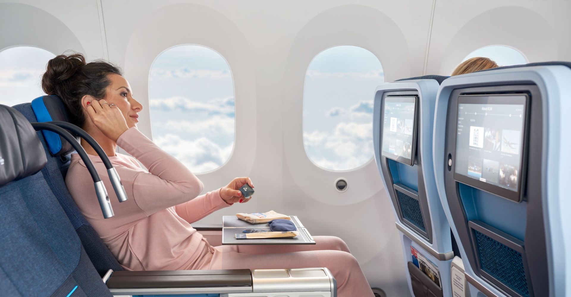 Komfortowa podróż w klasie Premium Comfort linii KLM – pasażerka relaksuje się na ergonomicznym fotelu, korzystając z zestawu podróżnego i rozrywki pokładowej. Nowoczesne wnętrze samolotu, większa przestrzeń i udogodnienia zapewniają wygodę na długich lotach. Sprawdź zalety podróży w klasie Premium Comfort KLM.