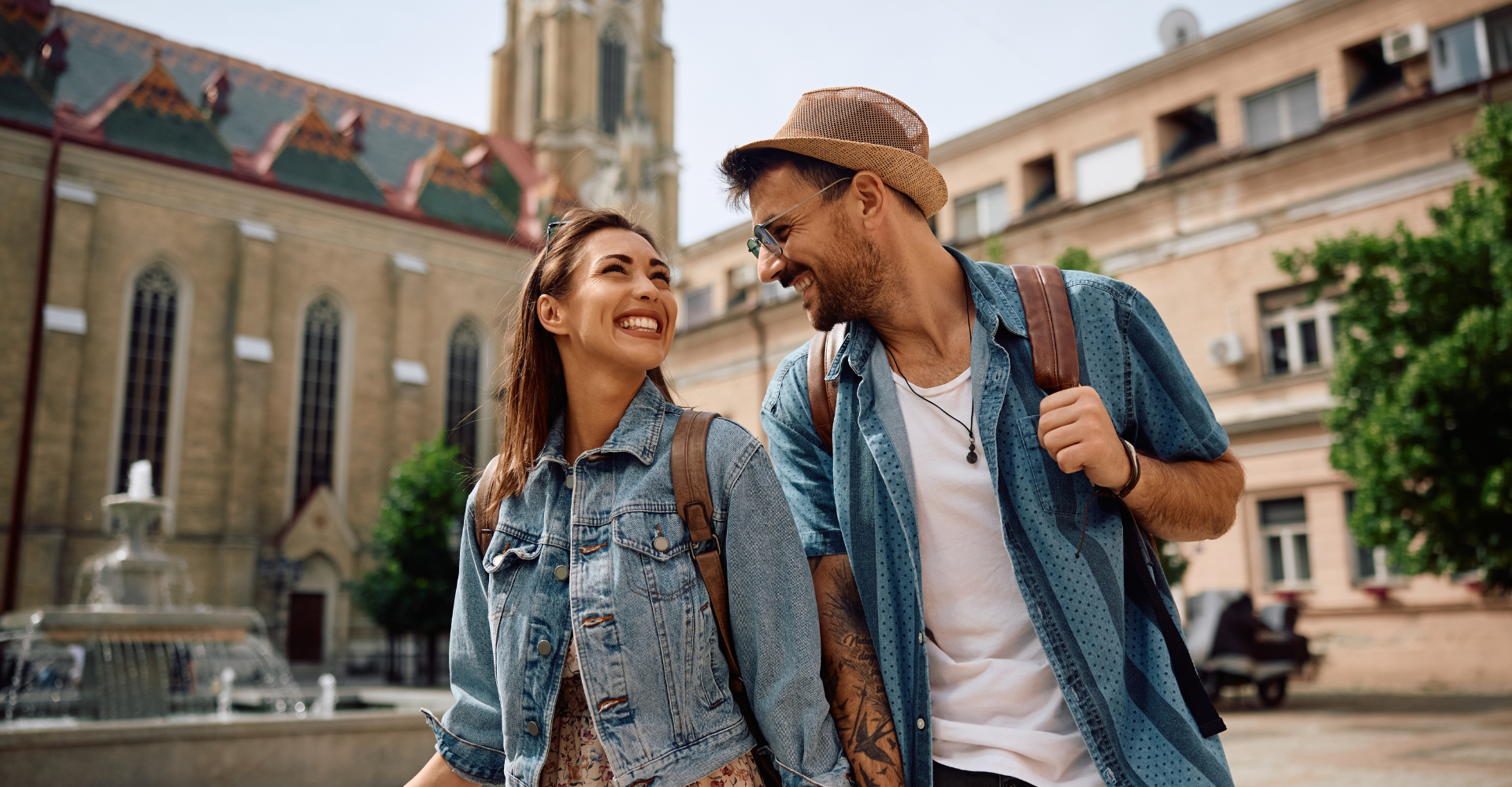 city break - Uśmiechnięta para turystów spacerująca po starym mieście w słoneczny dzień, w tle zabytkowy kościół i fontanna.