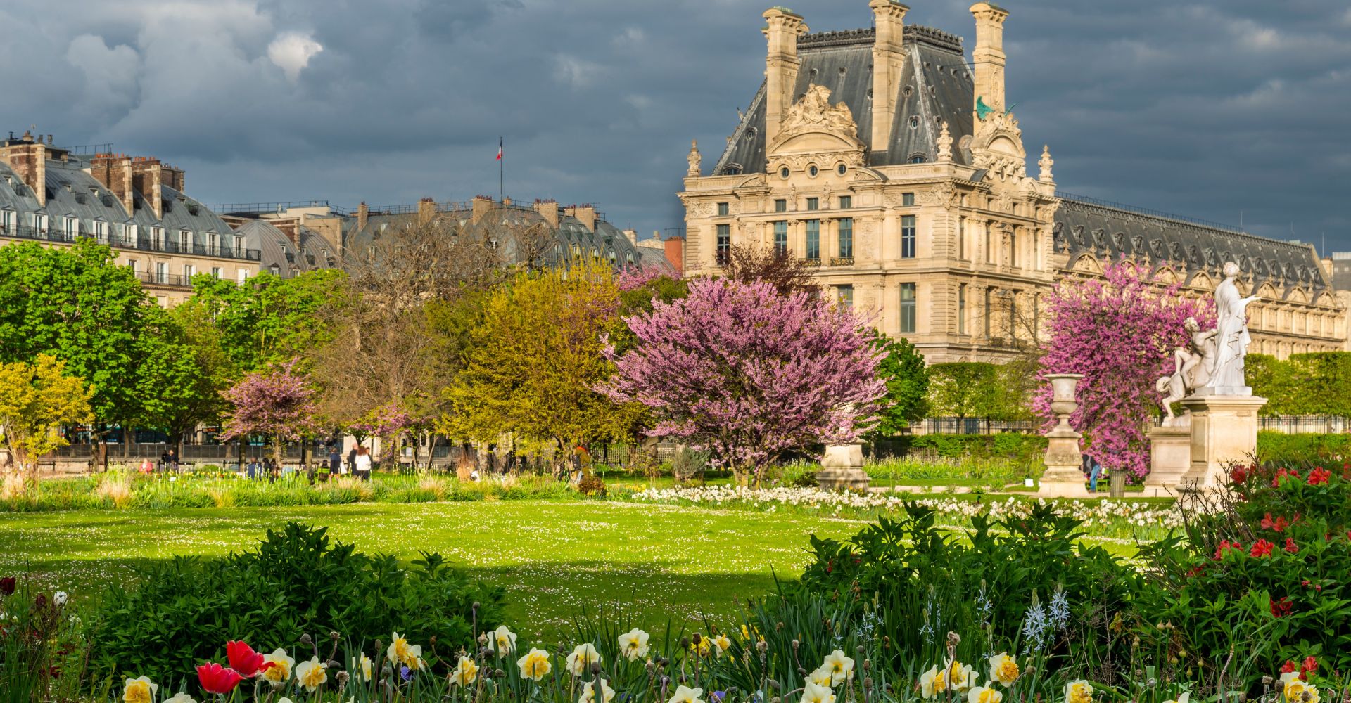 fragment Luwru w Paryżu. Budynek widoczny w tle to Pavillon de Flore – jedno ze skrzydeł kompleksu Luwru, który znajduje się przy Ogrodach Tuileries (Jardin des Tuileries), które widzisz na pierwszym planie z pięknie kwitnącymi drzewami i kwiatami. Shutterstock