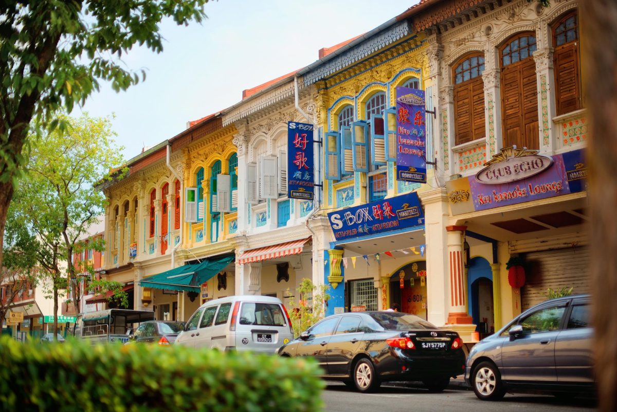 Singapur, Chinatown. Scena uliczna przedstawiająca rząd kolorowych domów w stylu Peranakan z ozdobnymi fasadami. Wzdłuż ulicy zaparkowane są samochody, a chodnik otaczają drzewa. Na budynkach widoczne są znaki w języku chińskim i angielskim, wskazujące na działalność takich firm jak salon karaoke.