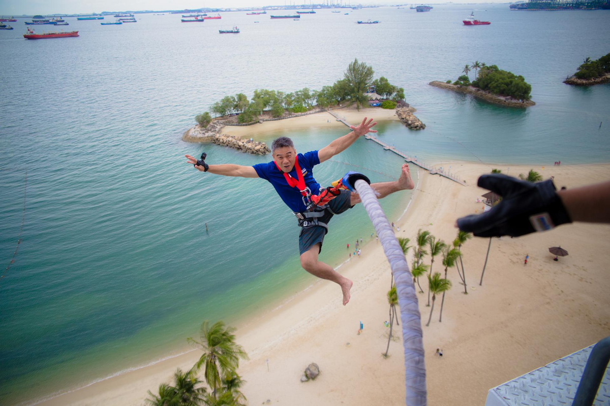 Daredevil Beach Bungee Turquoise Islands, Skypark Sentosa autorstwa AJ Hackett
Mężczyzna zawieszony w powietrzu na linie bungee, z rozpostartymi ramionami. Ma na sobie niebieską koszulkę, szare szorty i sprzęt zabezpieczający. W tle widoczna jest plaża z palmami, turkusowa woda i wyspy w oddali. Na horyzoncie widać kilka statków. Po prawej stronie częściowo widoczna jest ręka w rękawiczce.