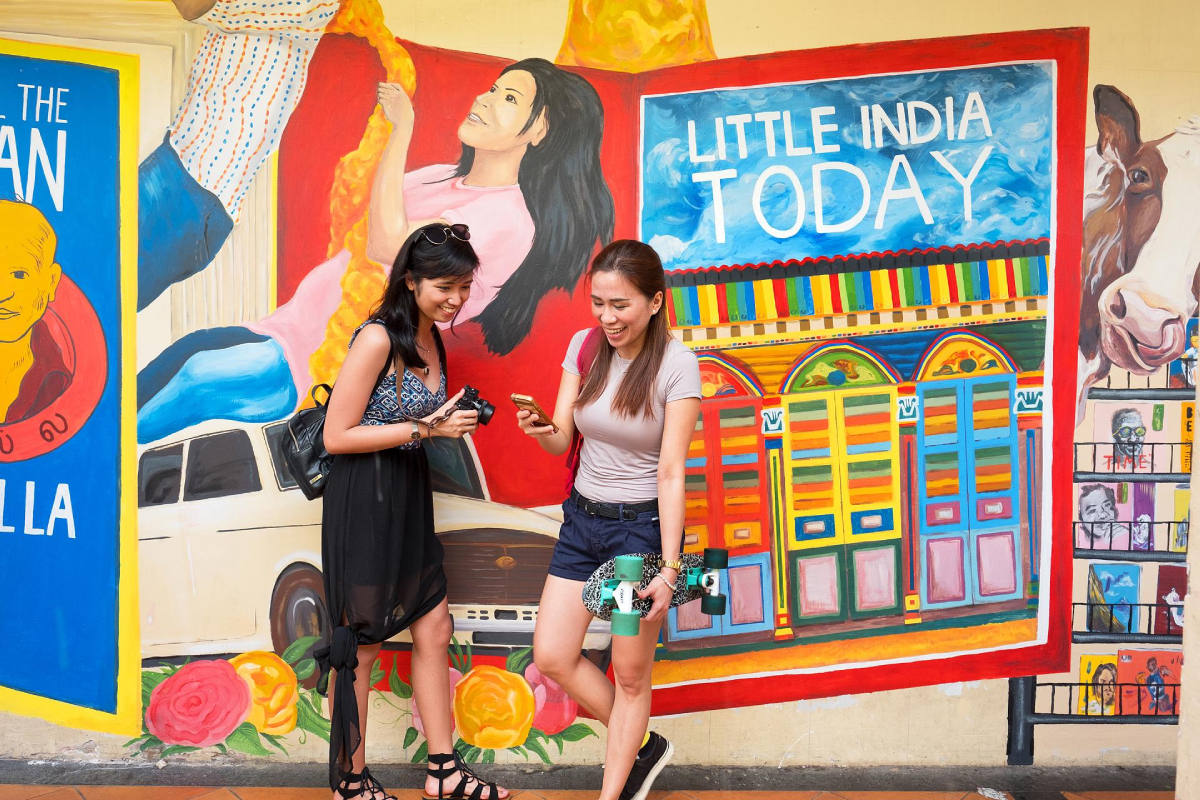 Singapur. Dwie kobiety stoją przed kolorowym muralem w dzielnicy Little India. Jedna z nich trzyma deskorolkę i patrzy na telefon, a druga trzyma aparat fotograficzny. Mural przedstawia różne obrazy, w tym portret kobiety, krowę i napis „Little India Today”.