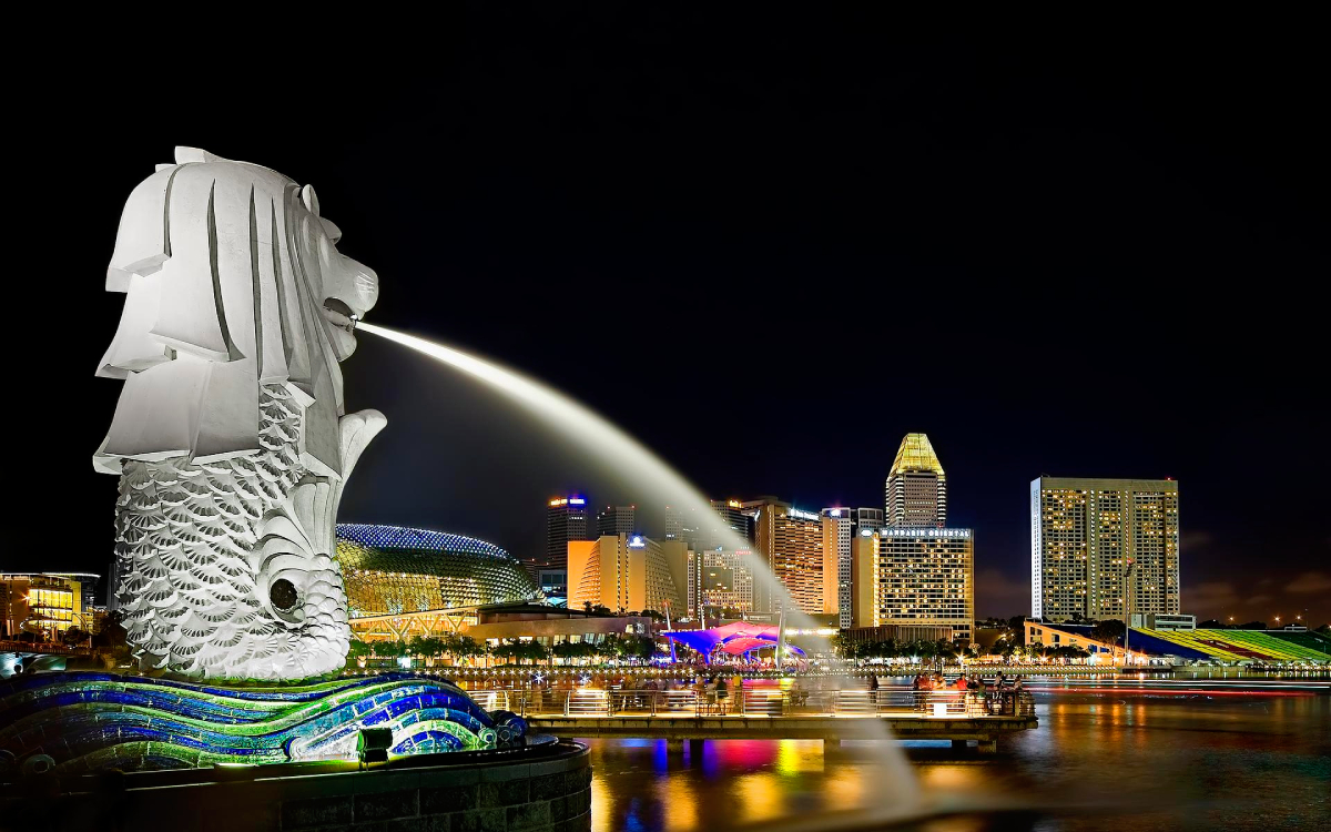 Nocna fontanna lwa na tle panoramy miasta, park Merlion
Centralnym punktem jest posąg Merlion, mitycznego stworzenia z głową lwa i ciałem ryby. Fontanna tryska wodą w kierunku nocnej panoramy miasta. W tle widoczne są budynki z oświetlonymi oknami i światłami.