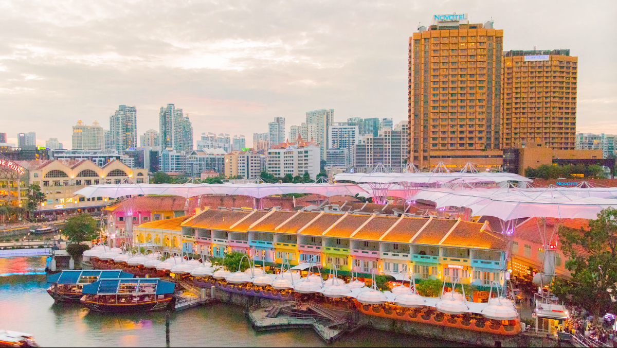 Riverside City Hotel Łodzie Panorama miasta, Clarke Quay
Widok z góry ukazuje tętniącą życiem panoramę miasta z rzeką otoczoną kolorowymi budynkami. Łodzie cumują wzdłuż brzegu, a w tle wznosi się duży hotel. Niebo jest zachmurzone.