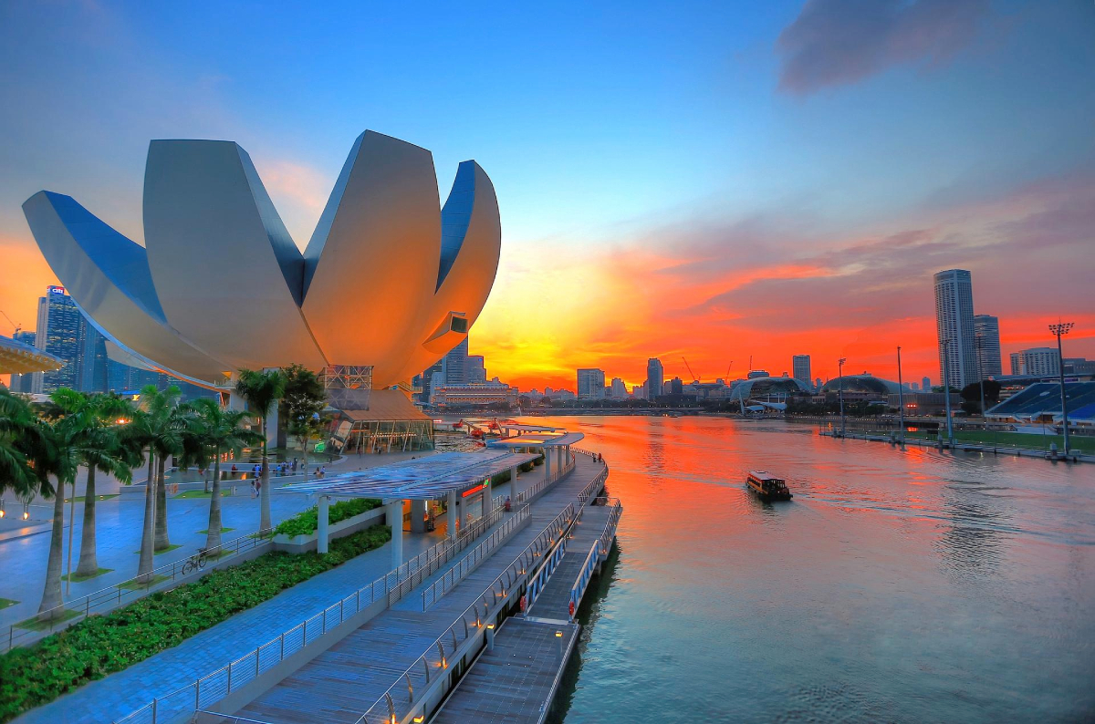 Singapur. Odbicie zachodu słońca w wodzie Budynek w kształcie kwiatu, Muzeum ArtScience
Architektoniczna konstrukcja przypominająca kwiat stanowi centralny punkt kompozycji na tle intensywnego zachodu słońca. Woda odbija kolory nieba, a na jej powierzchni widoczna jest łódź. Palmy otaczają chodnik, a budynki tworzą odległą linię horyzontu.