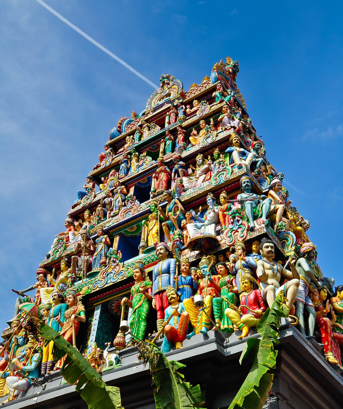 Singapur Sri Mariamman Temple. Kolorowa świątynia z wieloma szczegółowymi posągami ludzi i postaci. Świątynia ma trójkątny kształt, z wieloma rzędami posągów. Niebo jest błękitne z białym smugą kondensacyjną. Na pierwszym planie widoczne są zielone liście.
