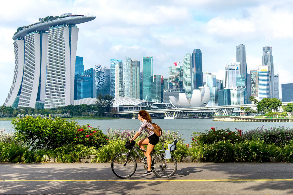 Singapur. Kobieta jedzie rowerem ścieżką, a w tle widać panoramę miasta. Wyraźnie widać hotel Marina Bay Sands. Ścieżka jest otoczona zielenią, a w wodzie odbija się panorama budynków. Niebo jest błękitne, z niewielką ilością chmur.