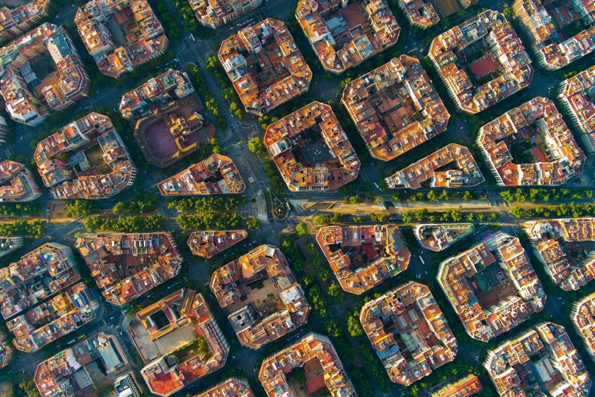 Zdjęcie przedstawia widok z lotu ptaka na dzielnicę Eixample w Barcelonie. Charakterystyczne ośmiokątne skrzyżowania i ścięte narożniki budynków tworzą regularną, geometryczną siatkę ulic. Widoczne są zielone aleje, które przecinają dzielnicę, otoczone harmonijnie rozmieszczonymi budynkami mieszkalnymi. Całość układu urbanistycznego ukazuje niezwykłą precyzję planowania miasta i podkreśla unikalny styl tej części Barcelony.