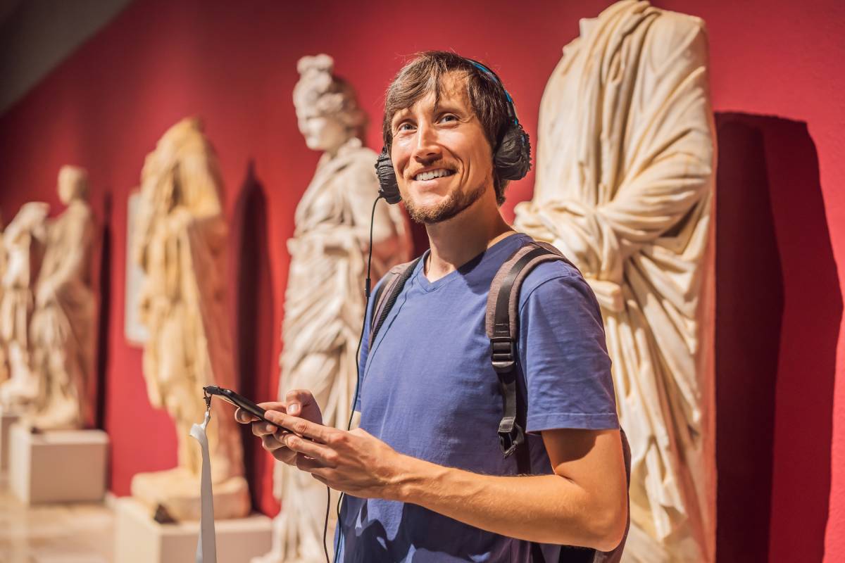 Uśmiechnięty mężczyzna z audioprzewodnikiem i smartfonem zwiedzający wystawę rzeźb w muzeum.