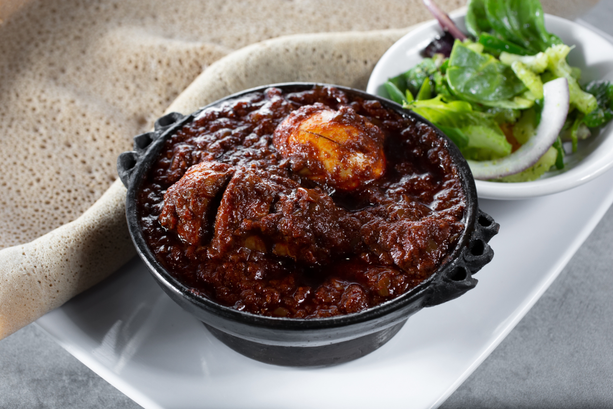 Etiopskie danie Doro Wat, gulasz z kurczaka i jajek w ostrym sosie berbere, podane w ciemnej misce z tradycyjnym chlebem injera i świeżą zieloną sałatką obok.