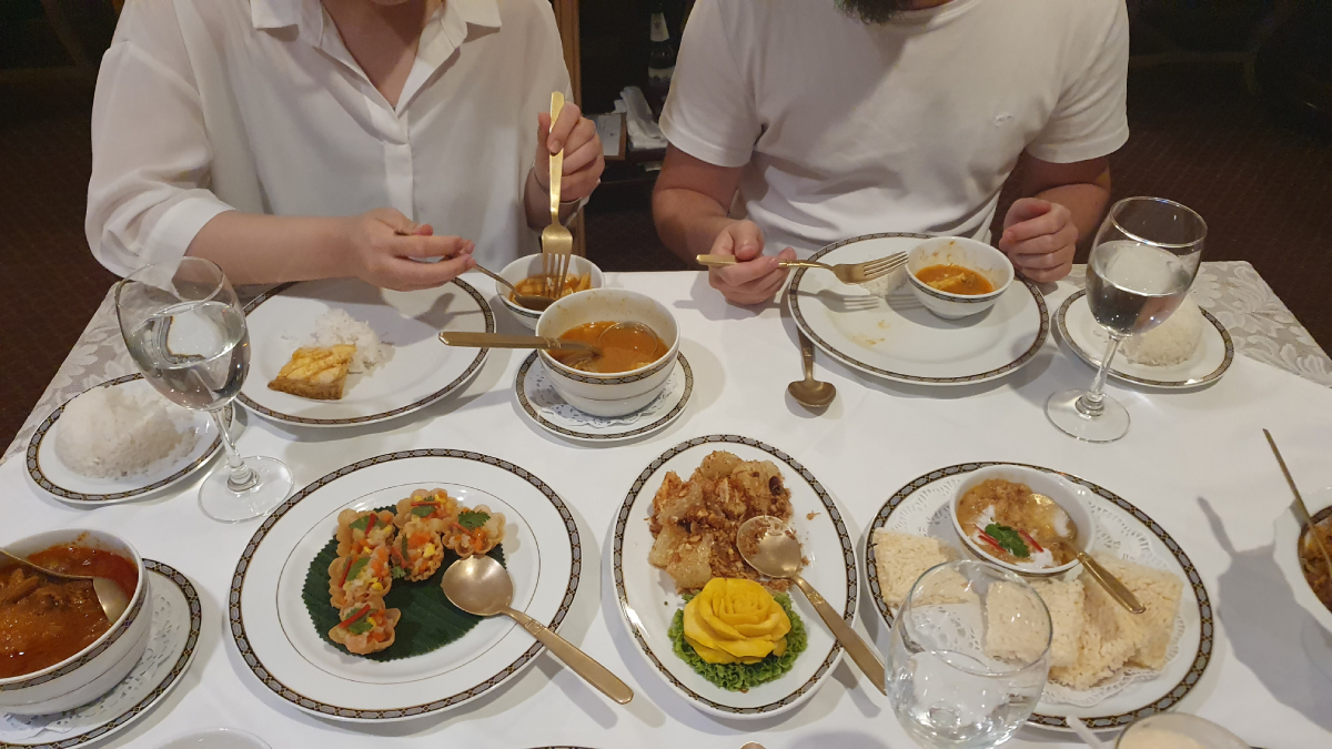 Zdjęcie stołu w tajskiej restauracji, zastawionego różnorodnymi potrawami, w tym zupą, daniami z ryżu i curry, przy którym siedzą dwie osoby jedzące posiłek.