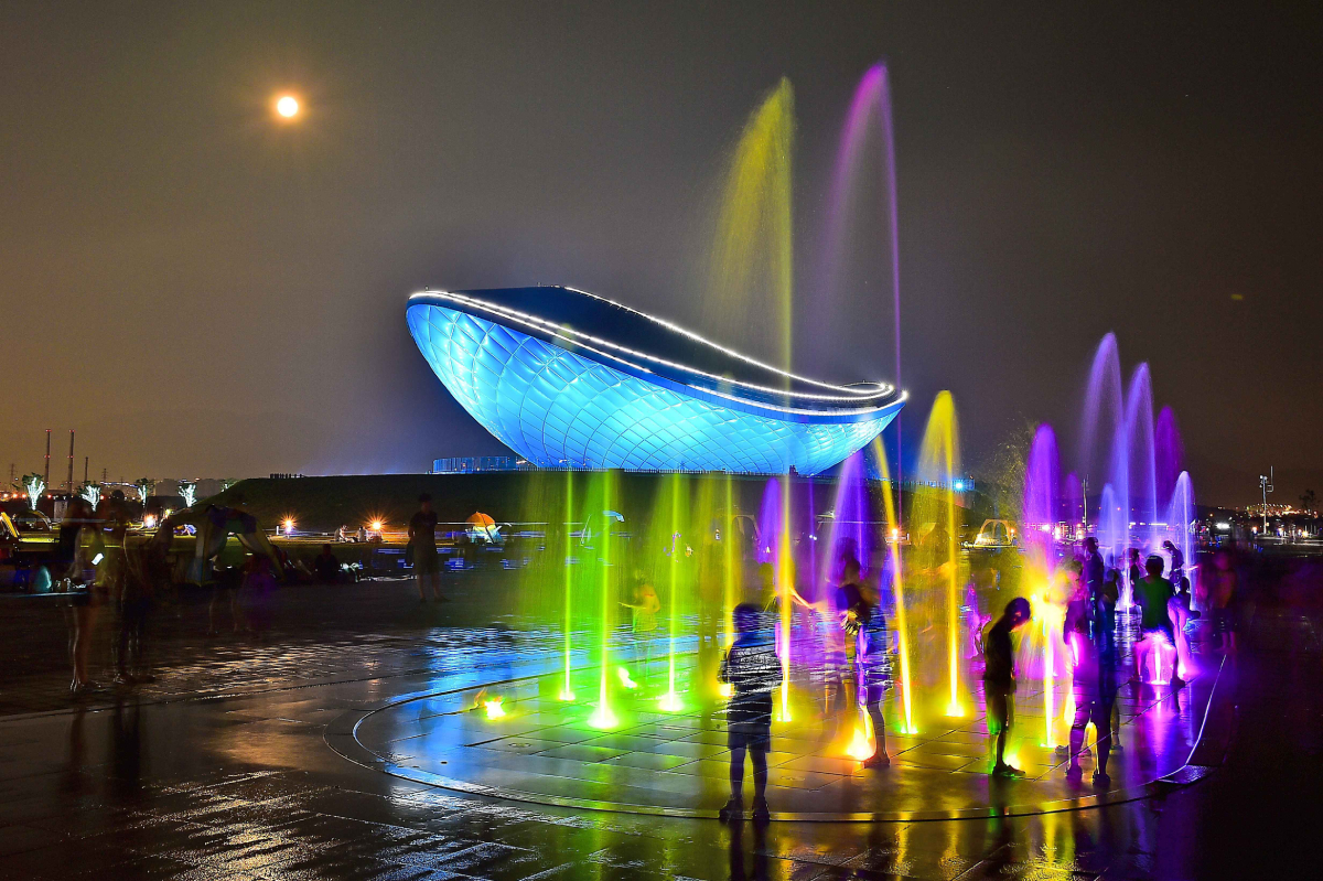 Nocna scena z podświetlaną fontanną i futurystycznym budynkiem Expo w Yeosu, Korea Południowa; dzieci bawią się wśród kolorowych strumieni wody, a w tle świeci księżyc.