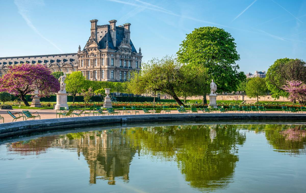 Wiosenny widok na Ogrody Tuileries z fontanną, rzeźbami i bujną zielenią, w tle pawilon Luwru.