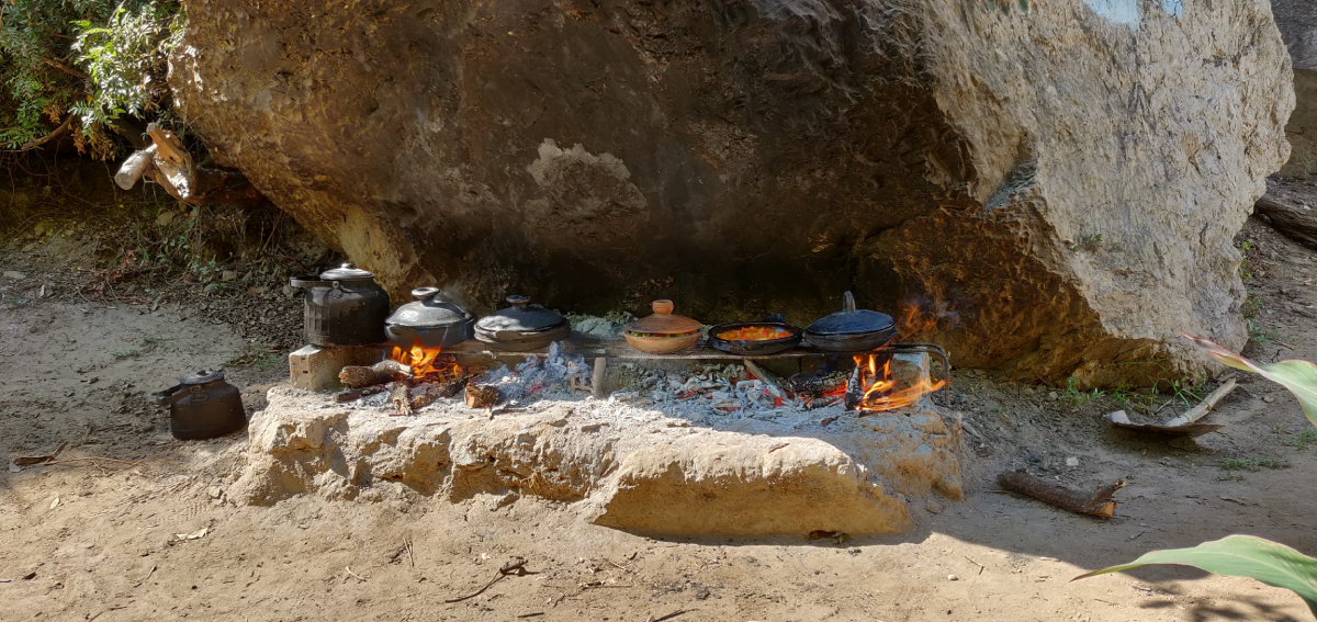 Tradycyjne marokańskie naczynia tagine i garnki gotujące się na otwartym ogniu pod naturalną skalną osłoną, z dymem unoszącym się z paleniska, podkreślające autentyczność wiejskiego gotowania.