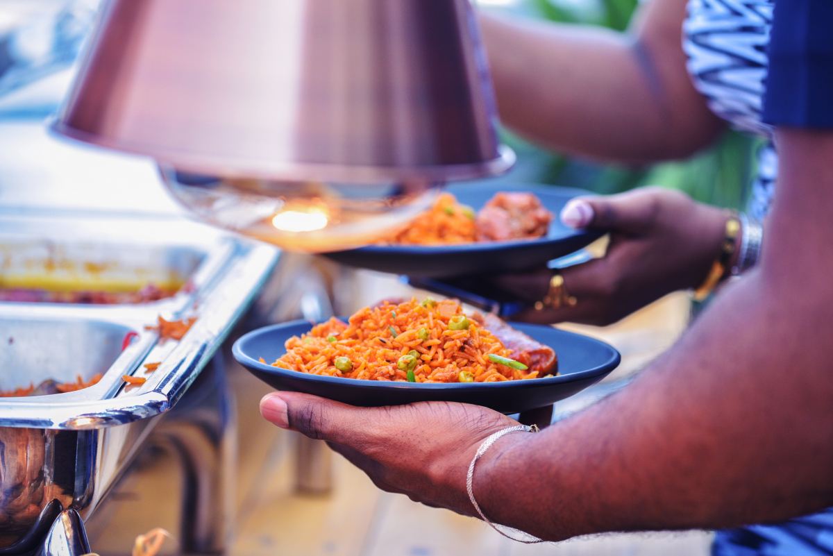 Dłonie trzymające talerze z nigeryjskim ryżem Jollof, z dodatkiem grochu i kawałków mięsa wołowego (Pomo), podawanym w formie bufetu.