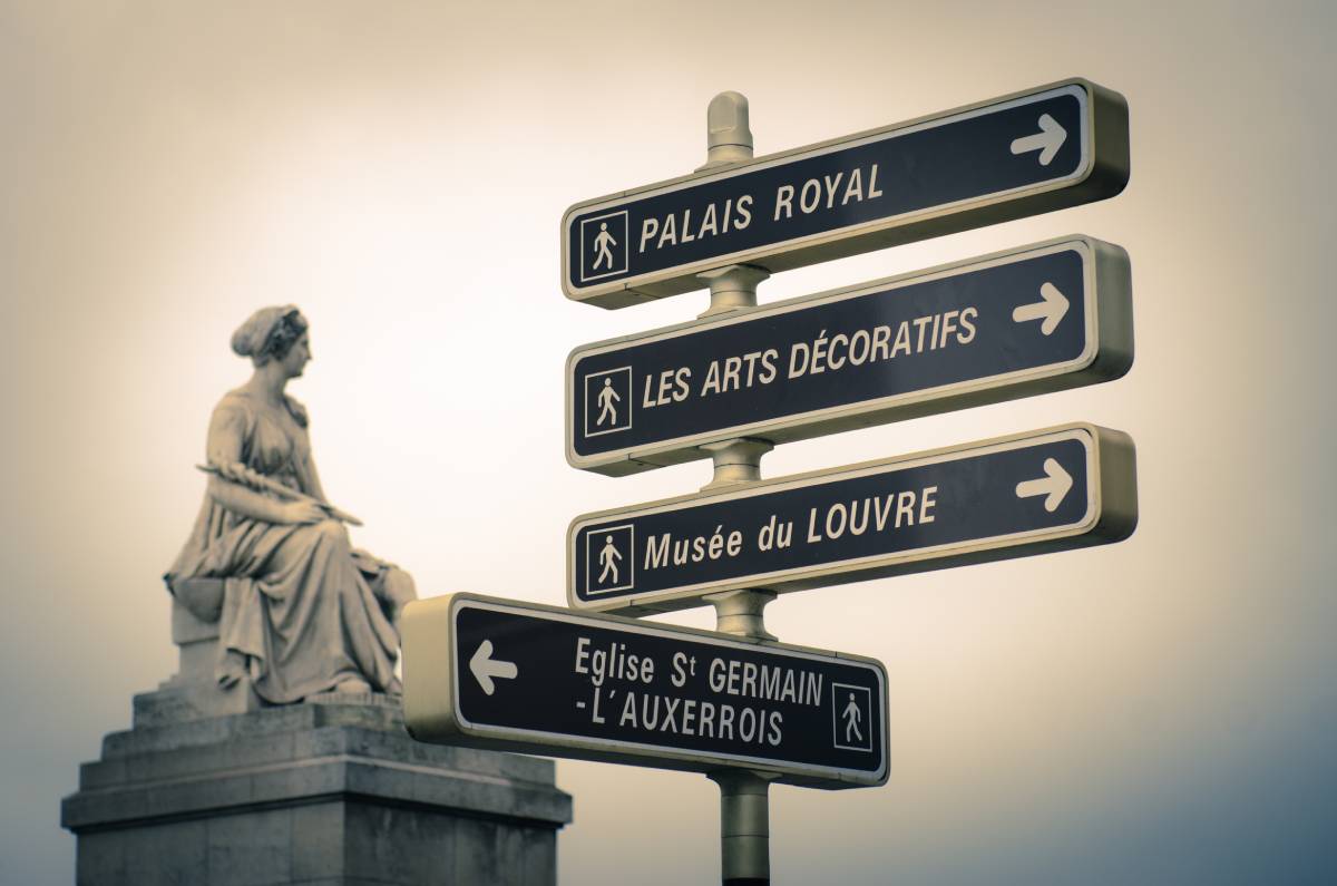 Znak kierunkowy w Paryżu wskazujący drogę do Musée du Louvre, Palais Royal, Les Arts Décoratifs i kościoła Saint-Germain-l'Auxerrois, w tle klasyczna rzeźba kobiety.
