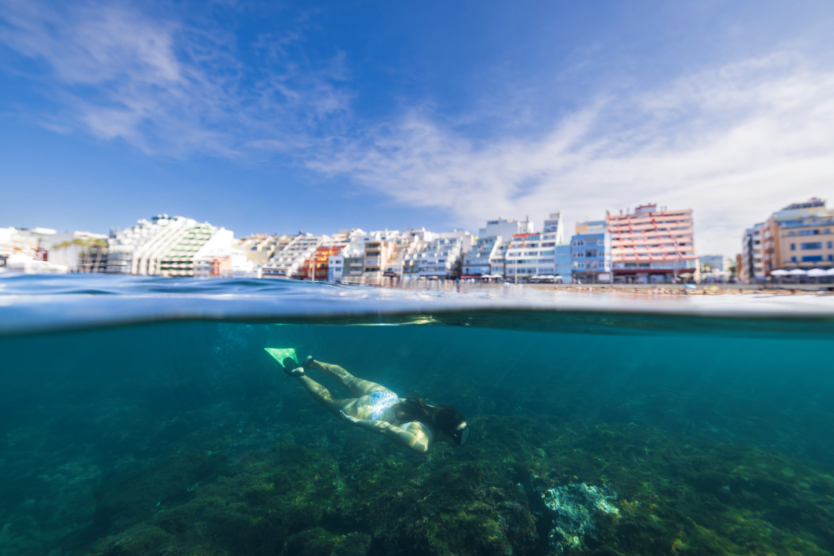 Snorkeler pływający w przejrzystej wodzie przy Playa de Las Canteras w Las Palmas de Gran Canaria, z panoramą miejską w tle.