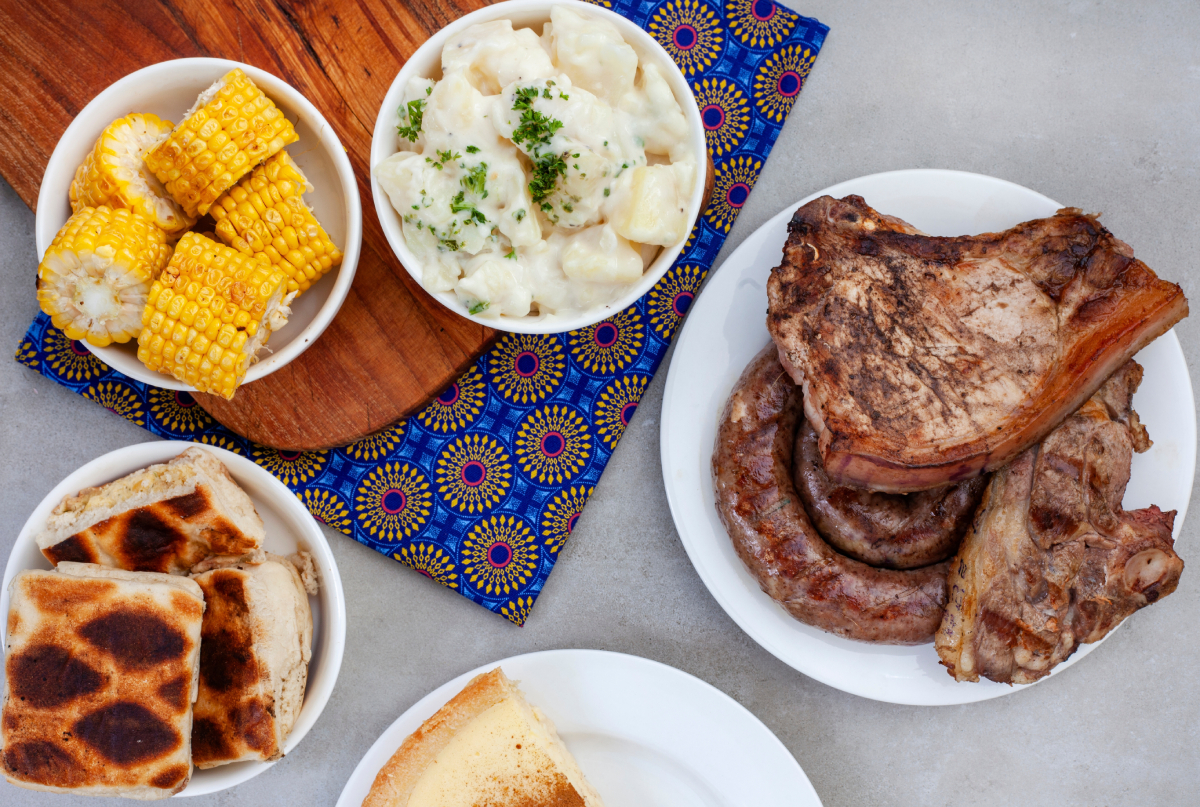 Stół zastawiony tradycyjnymi potrawami z południowoafrykańskiego Braai Day (Dzień Dziedzictwa), obejmujący grillowane mięso (żeberka i kiełbaski boerewors), kolby kukurydzy, sałatkę ziemniaczaną, tosty oraz ciasto, wszystko ułożone na tle tkaniny Shwe-Shwe.