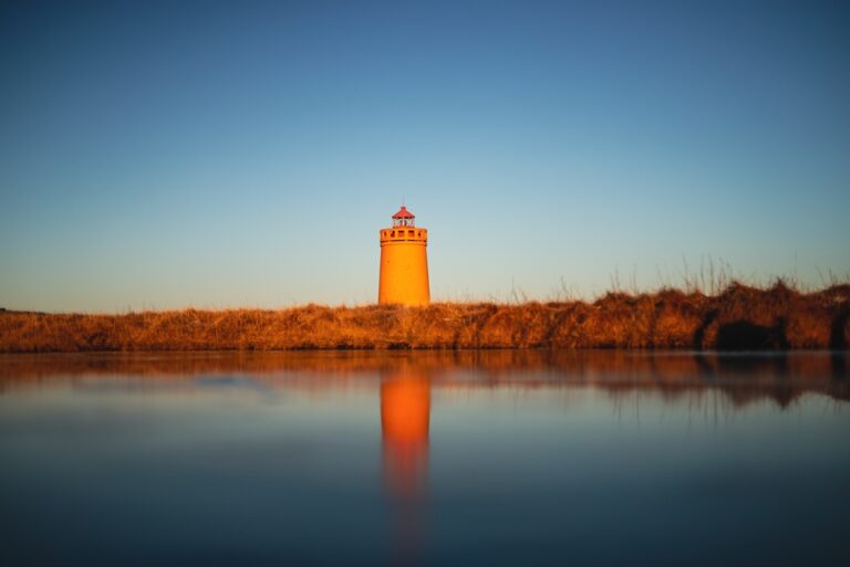 Pomarańczowa latarnia morska w Keflavíku na Islandii, odbijająca się w spokojnej tafli wody na tle zimowego krajobrazu