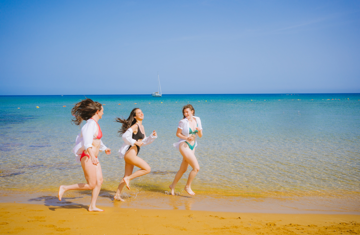 Trzy turystki biegnące brzegiem morza na piaszczystej plaży Ramla Bay na wyspie Gozo, Malta.