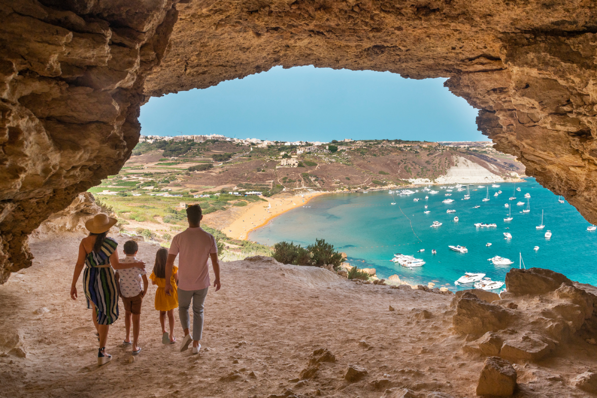 Rodzina podziwiająca panoramę Ramla Bay na Gozo z wejścia do groty skalnej, z widokiem na plażę i zatokę z białymi jachtami.