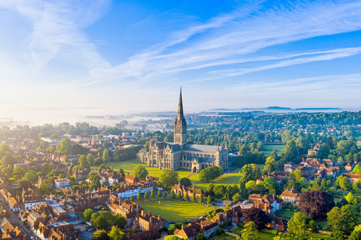 Panorama angielskiego miasta Salisbury z lotu ptaka o poranku, z dominującą na pierwszym planie monumentalną gotycką katedrą otoczoną zielenią.