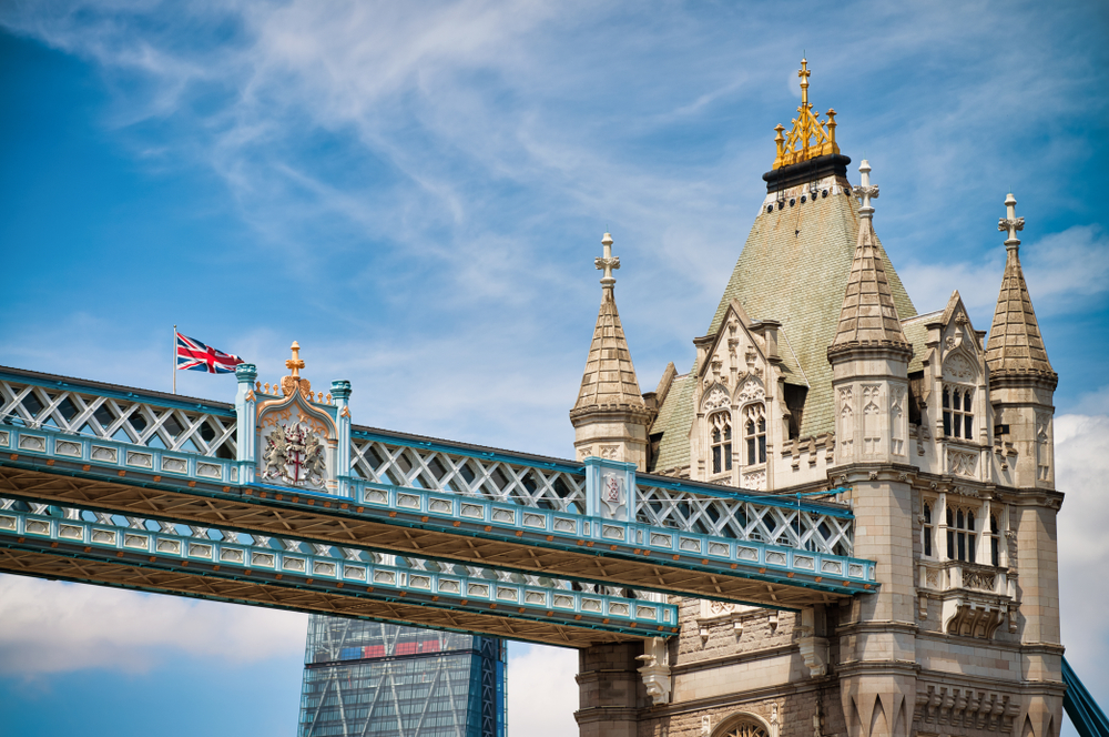 Zbliżenie na wieżę i górną kładkę mostu Tower Bridge w Londynie podczas letniego dnia, z widoczną brytyjską flagą na tle błękitnego nieba.