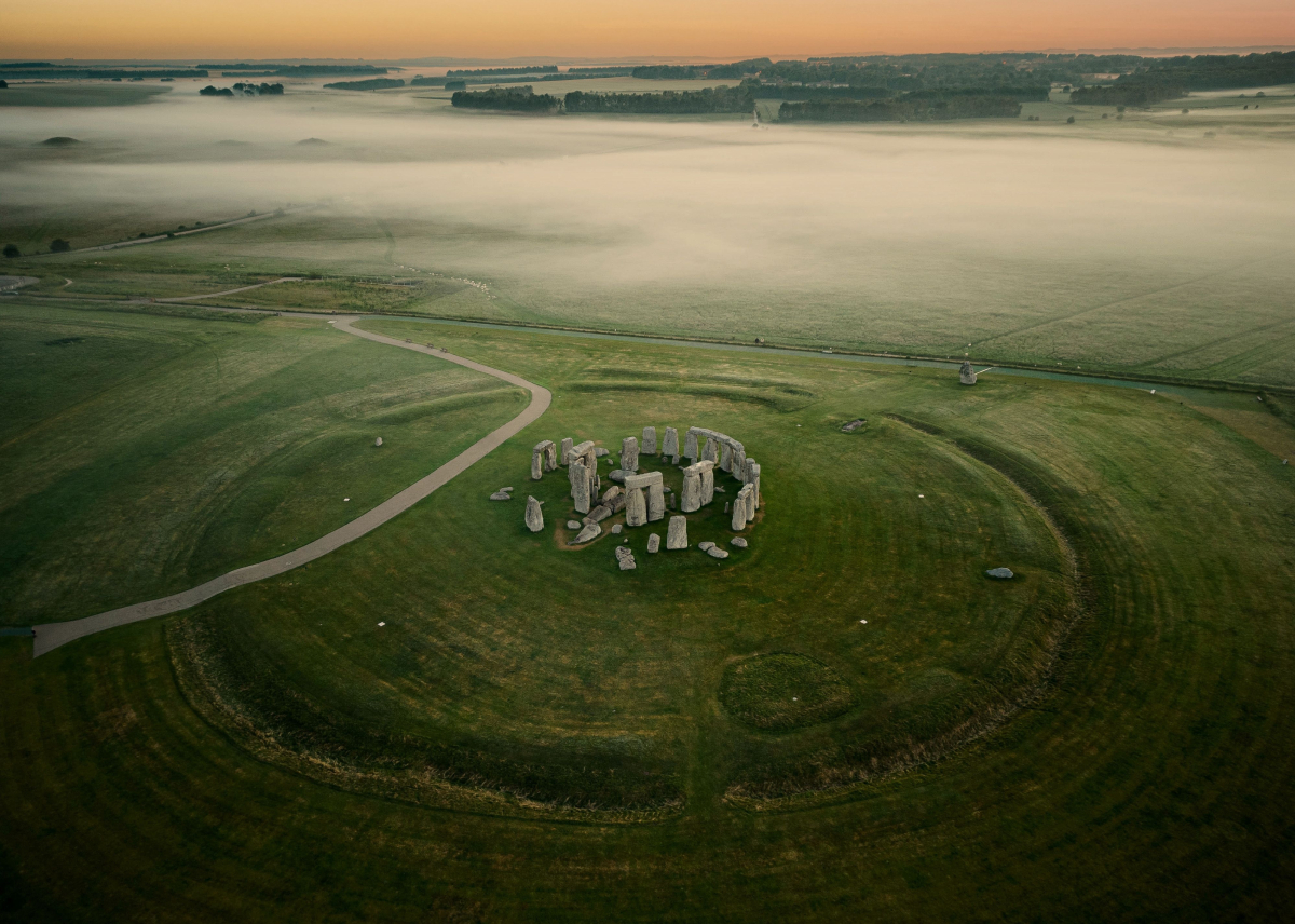 Widok z lotu ptaka na Stonehenge o poranku, otoczone mgłą i zielonymi polami, ukazujący tajemniczy układ kamiennych bloków.