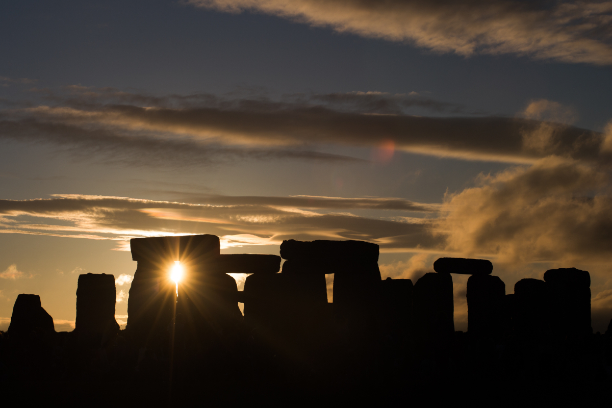Sylwetka kamieni Stonehenge uchwycona pod słońce o świcie, z promieniami światła przenikającymi przez szczelinę między kamieniami.