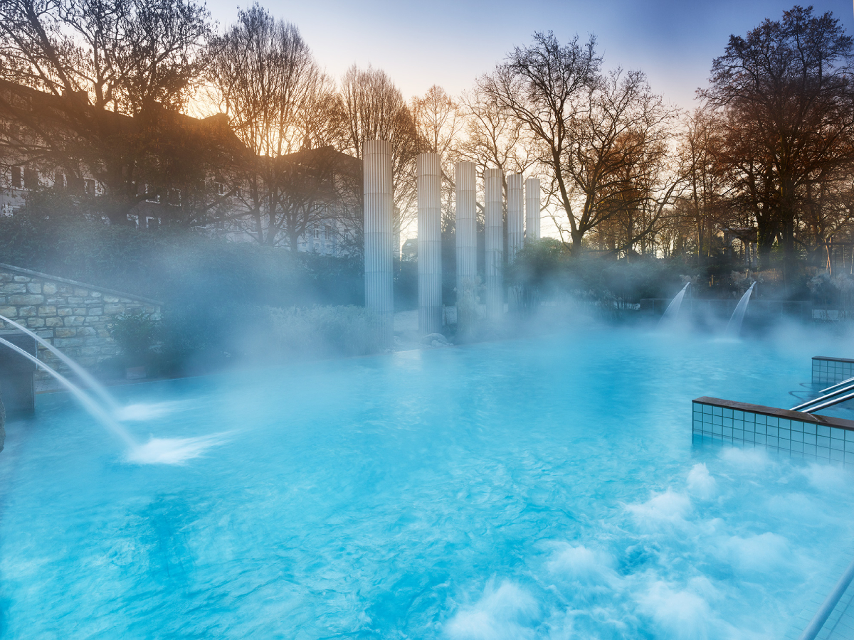 Zewnętrzny basen z turkusową wodą w termach Carolus Therme w Aachen, z widocznymi unoszącymi się kłębami pary. W tle znajdują się skalne ściany i drzewa, a woda wypływa z dysz wprost do basenu. Zdjęcie przedstawia idealne miejsce na relaks i odpoczynek.