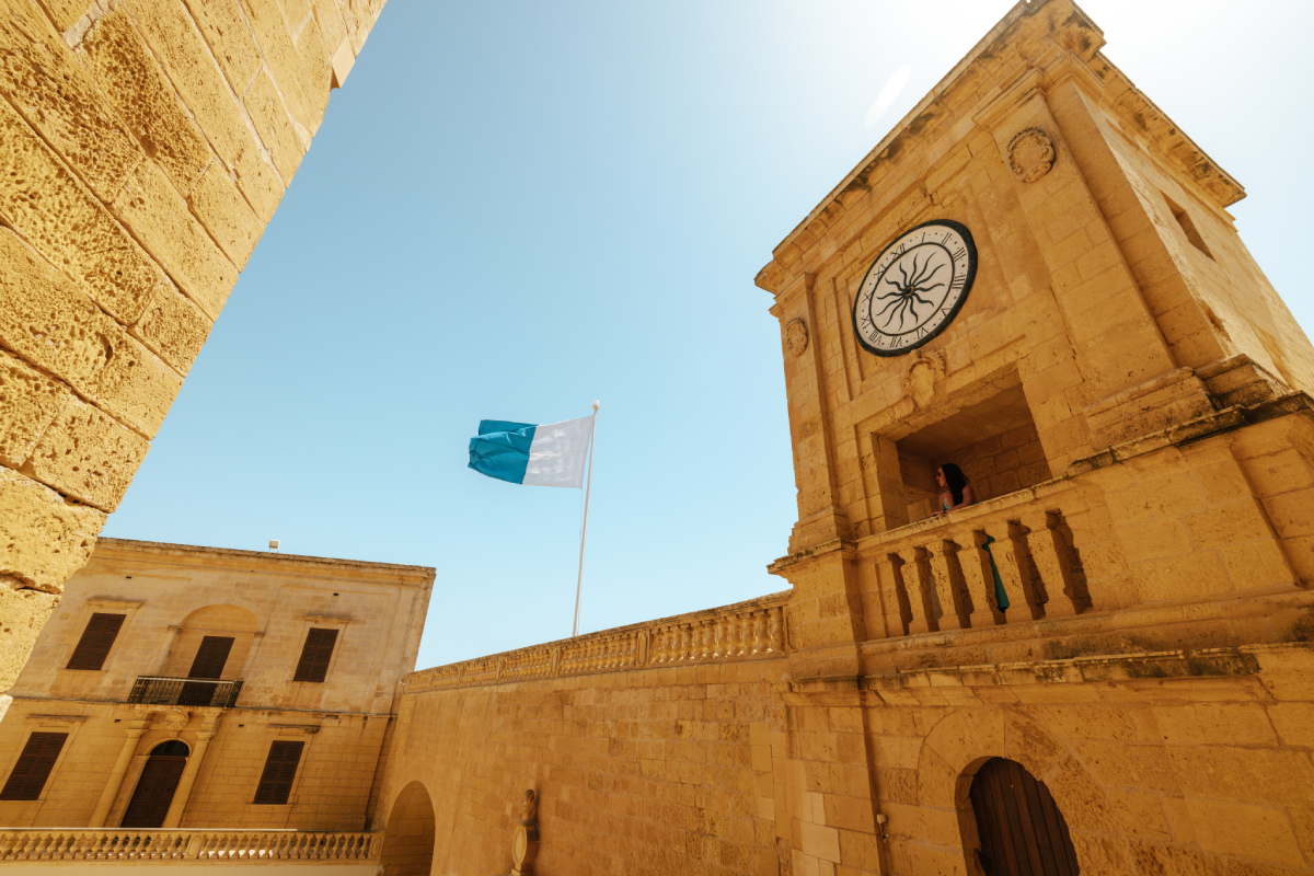 Wieża zegarowa Cytadeli w Victorii na wyspie Gozo, Malta, z powiewającą biało-niebieską flagą na tle błękitnego nieba.