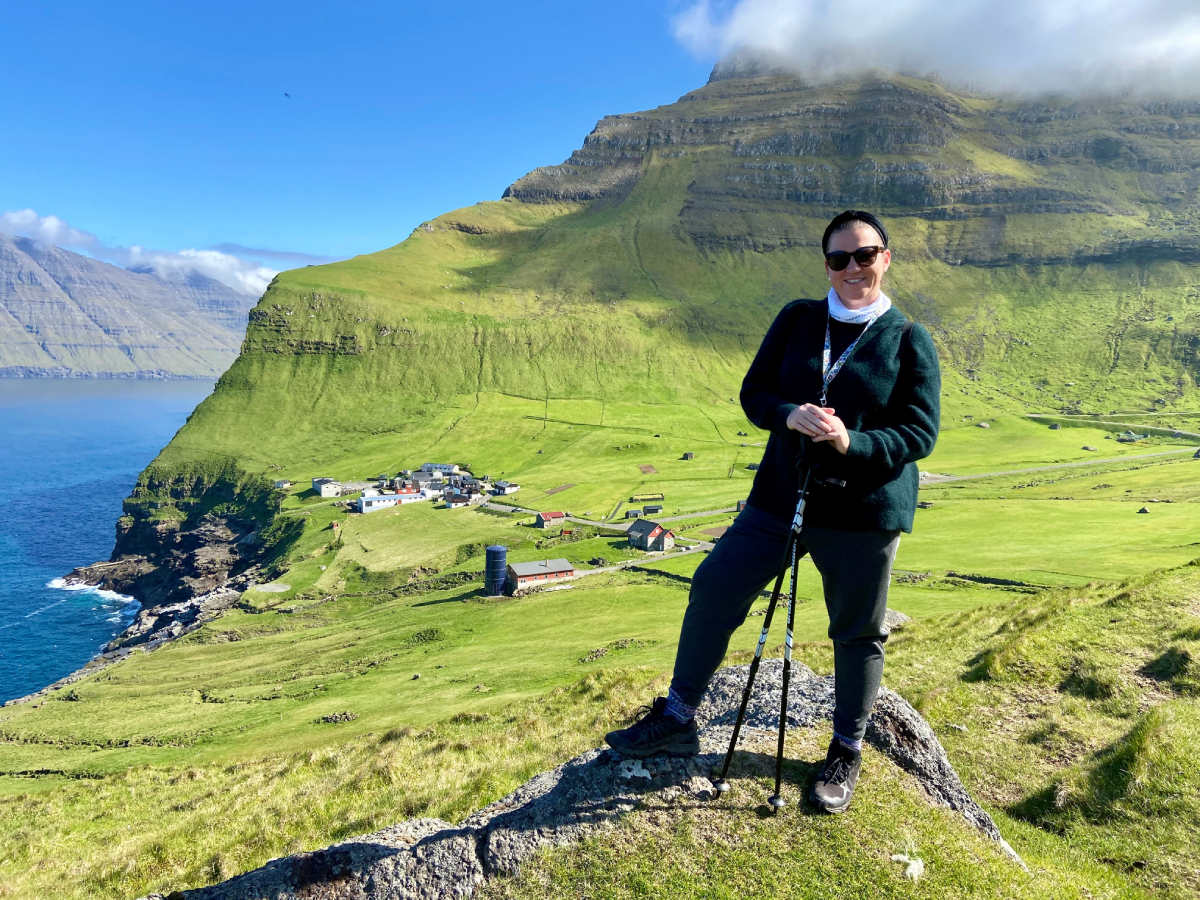 Kinga Eysturland podczas trekkingu na wyspie Kalsoy na Wyspach Owczych, z malowniczą wioską i stromymi klifami w tle